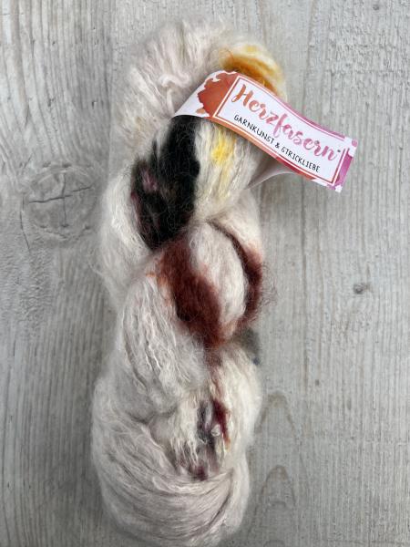 Ein heller Strang Suri Alpaka mit farbigen Sprenkeln in Schwarz, Gelb, Rotbraun und Pink, handgefärbt, liegend auf einem Holzbrett.